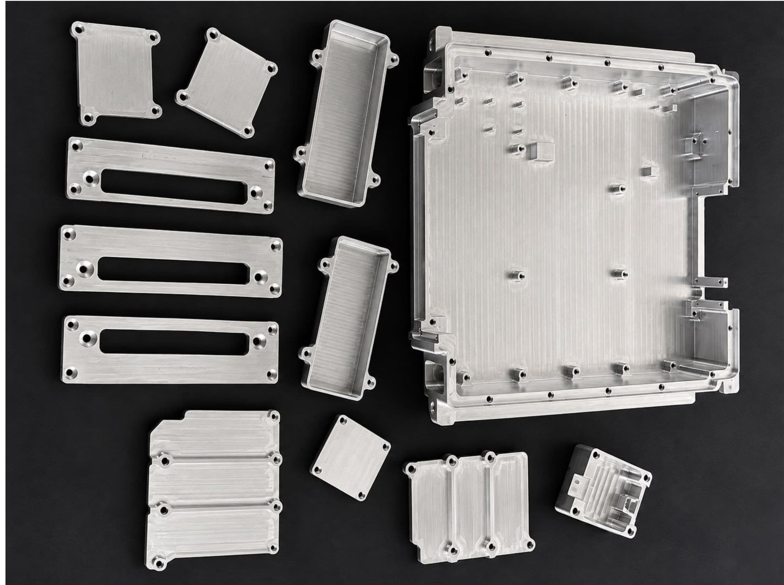 A machined enclosure base with matching lids, trays, slotted frames and small covers laid out on a dark surface.